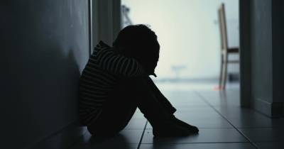 Sad child suffering from depression sitting alone in corridor feeling loneliness. Scared fearful small boy covering face in silhouette at home