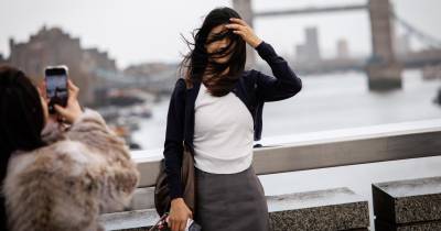 Vento forte afeta Reino Unido. Mulher tenta posar para uma foto, com a London Bridge ao fundo.